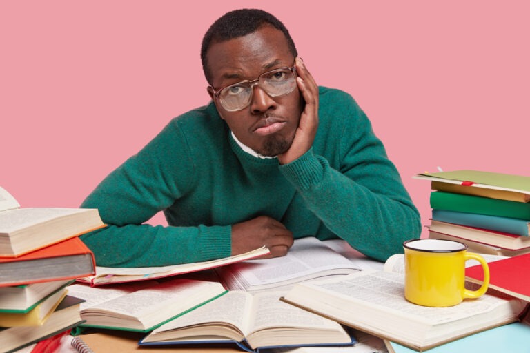 Upset dark skinned man has bored expression, keeps hands on cheek, wears spectacles and green sweater, surrounded with many books, drinks coffee, isolated over pink background. Ethnicity concept